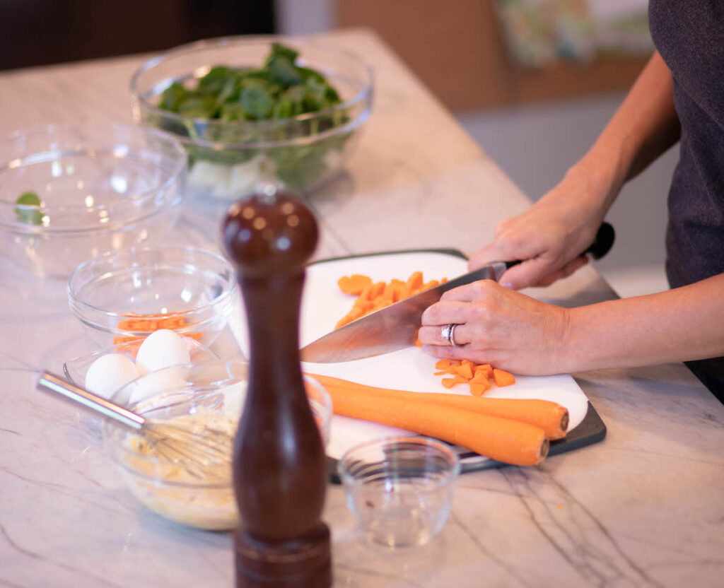 Hands cutting carrots on a cutting bowls with other food on counter for Freezer tips post.