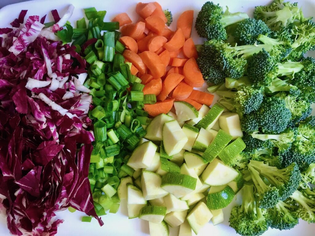 Red cabbage chopped, scallions sliced, carrots and zucchini diced, and broccoli florets all sitting next to each other on a cutting board.