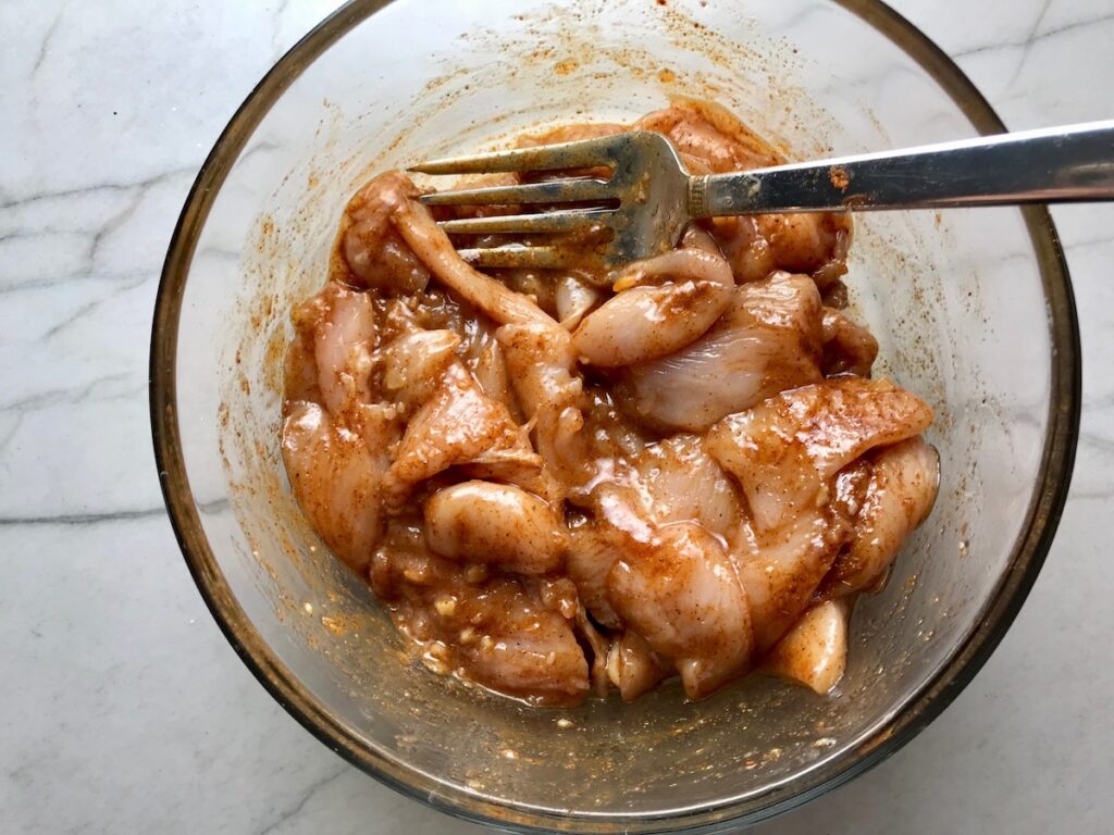 Raw Mexican Marinated chicken strips in a clear bowl with a fork.