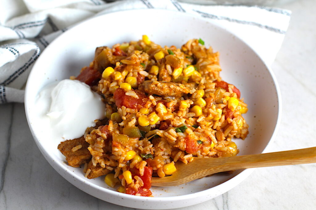 Mexican Marinated Chicken and Rice in a bowl with tomatoes, corn, scallions, and a dollop of sour cream on the side. Bamboo fork scooping inside the bowl and skillet in background.