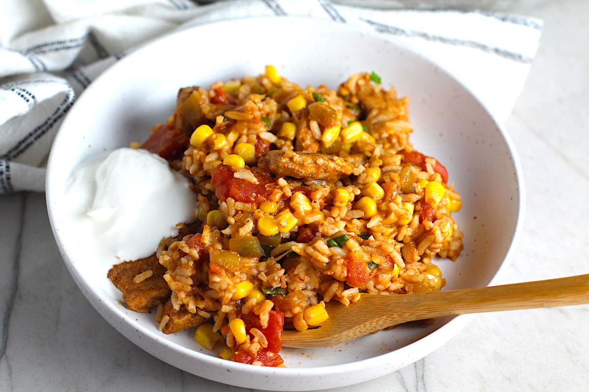 Mexican Marinated Chicken and Rice in a bowl with tomatoes, corn, scallions, and a dollop of sour cream on the side. Bamboo fork scooping inside the bowl and skillet in background.