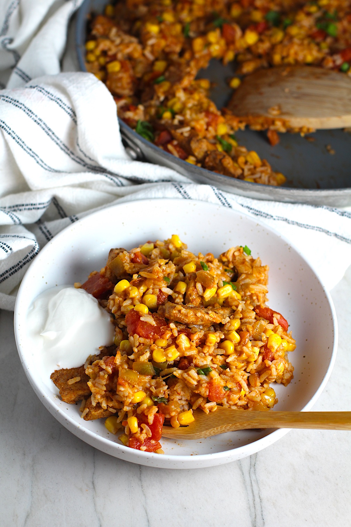 Mexican Marinated Chicken and Rice in a bowl with tomatoes, corn, scallions, and a dollop of sour cream on the side. Bamboo fork scooping inside the bowl and skillet in background..