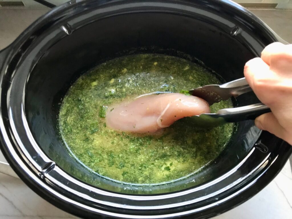 Tongs adding raw chicken breast to broth and herbs in crockpot for Ranch Chicken Crock Pot Pasta recipe. It uses fresh dill, parsley, and scallions is creamy, tangy, and utterly delicious! It's also so easy to make, so it's a great weeknight dinner.