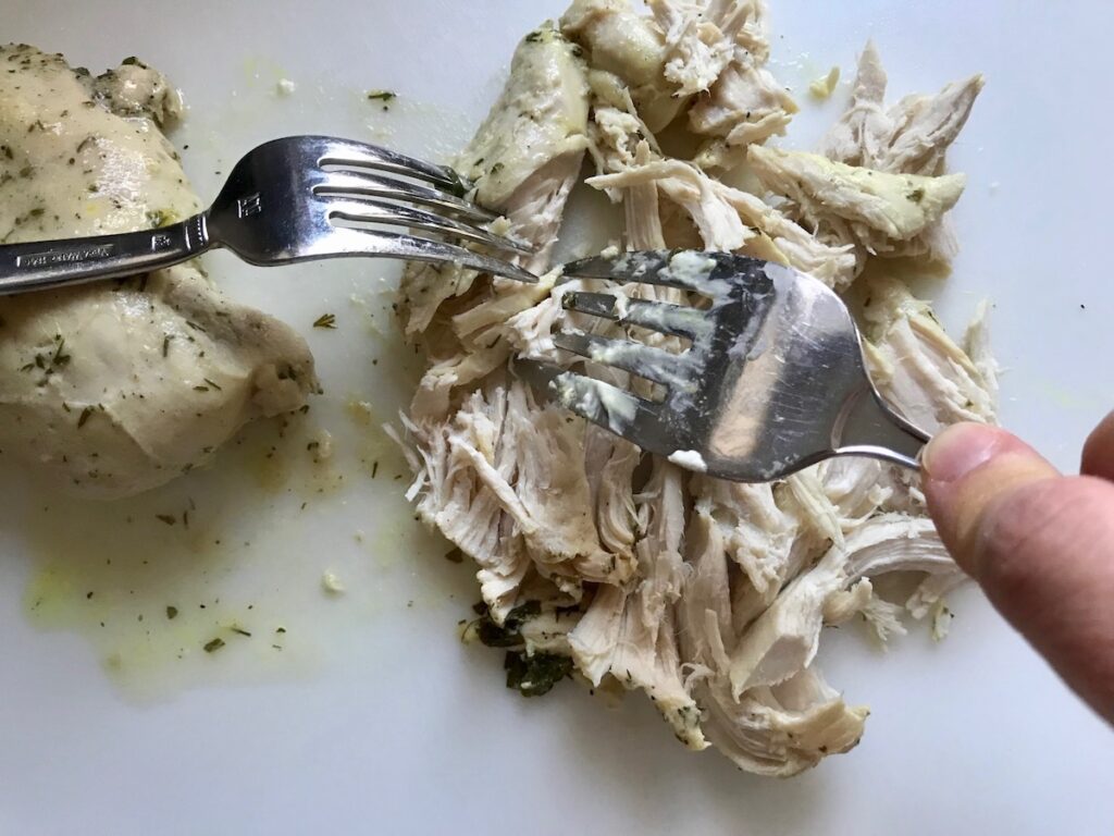 Two forks shredding cooked chicken breast for Ranch Chicken Crock Pot Pasta recipe. It uses fresh dill, parsley, and scallions is creamy, tangy, and utterly delicious! It's also so easy to make, so it's a great weeknight dinner.