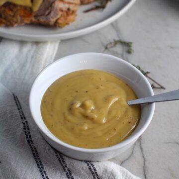 Thick brown Roast Pork Gravy recipe in a bowl with spoon with towel next to bowl and roast pork tenderloin in background. This Roast Pork Gravy Recipe is thick, creamy, and full of flavor.