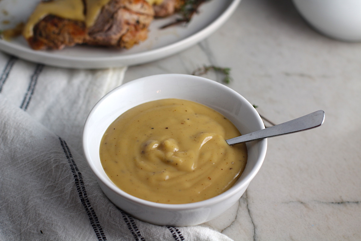 Thick brown Roast Pork Gravy recipe in a bowl with spoon with towel next to bowl and roast pork tenderloin in background. This Roast Pork Gravy Recipe is thick, creamy, and full of flavor.