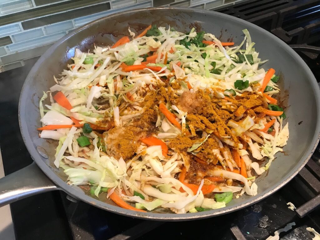Cabbage, carrots, spices and seasonings added to pan on stove for Shrimp Mei Fun Recipe, Singapore Noodles. It's an easy stir fry to make at home and tastes so much better than take out! With carrots, bean sprouts, curry powder, sesame oil, and more it's is a 30 minute weeknight dinner.