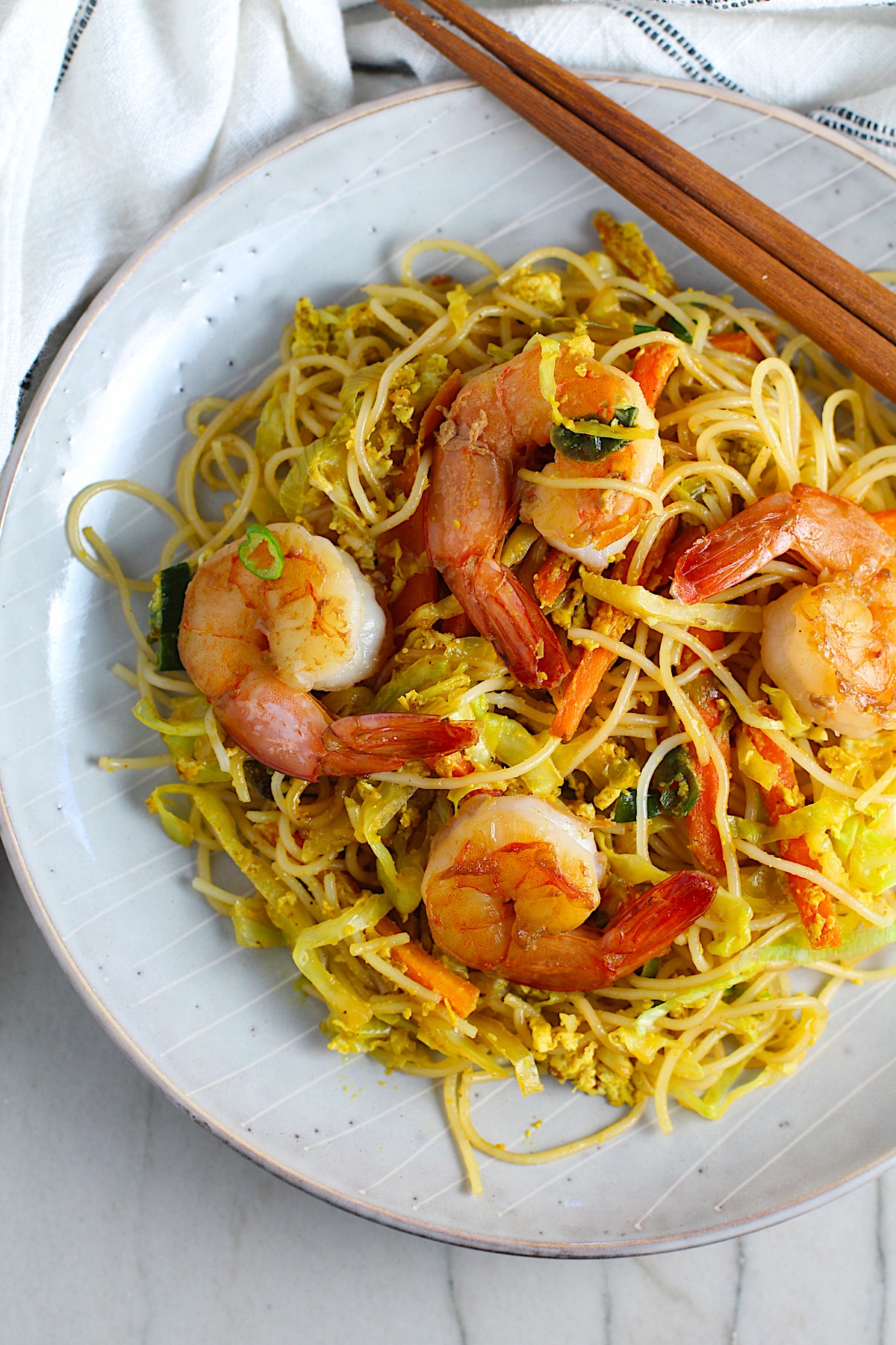Shrimp Mei Fun Recipe, Singapore Noodles, on a plate with brown wood chop sticks on counter with napkin. It's an easy stir fry to make at home and tastes so much better than take out! With carrots, bean sprouts, curry powder, sesame oil, and more it's is a 30 minute weeknight dinner.
