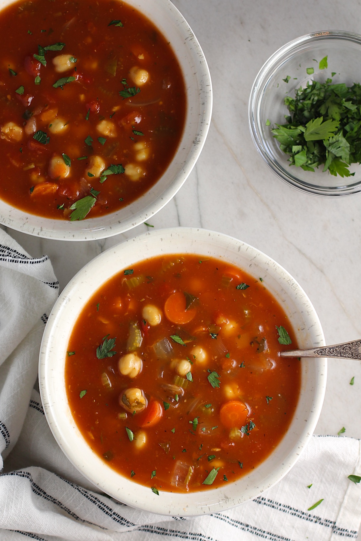 Two bowls of Vegetable Garbanzo Soup on a counter with a towel and a bowl of cilantro to the side.  It's slightly creamy and filled with flavor and texture! It's super easy to make ahead and enjoy during the week.