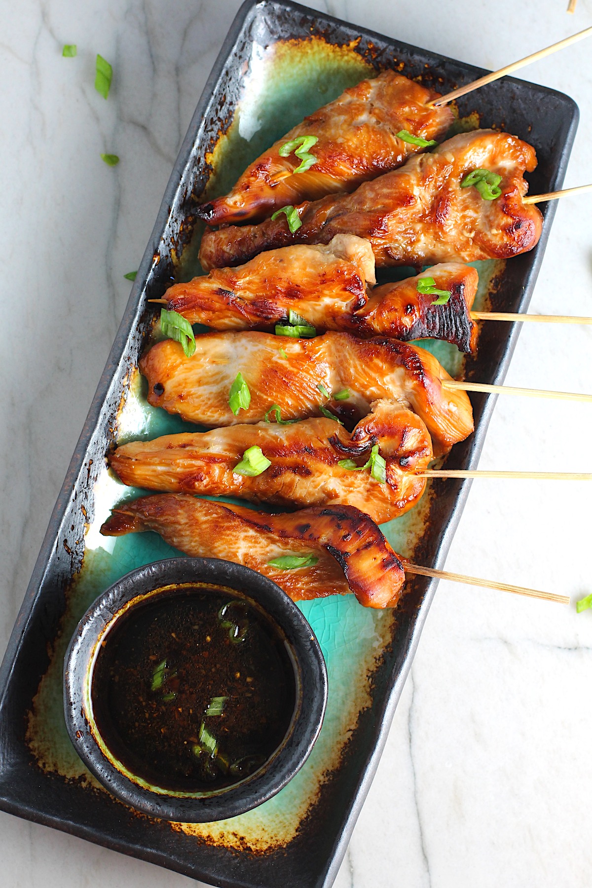 Chinese Chicken on a Stick Recipe on a rectangle platter with small bowl of dipping sauce and pan in background.