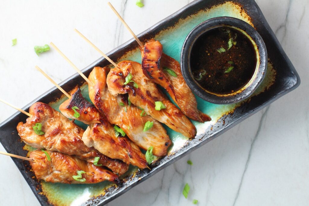 Chinese Chicken on a Stick Recipe on a rectangle platter with small bowl of dipping sauce and pan in background.