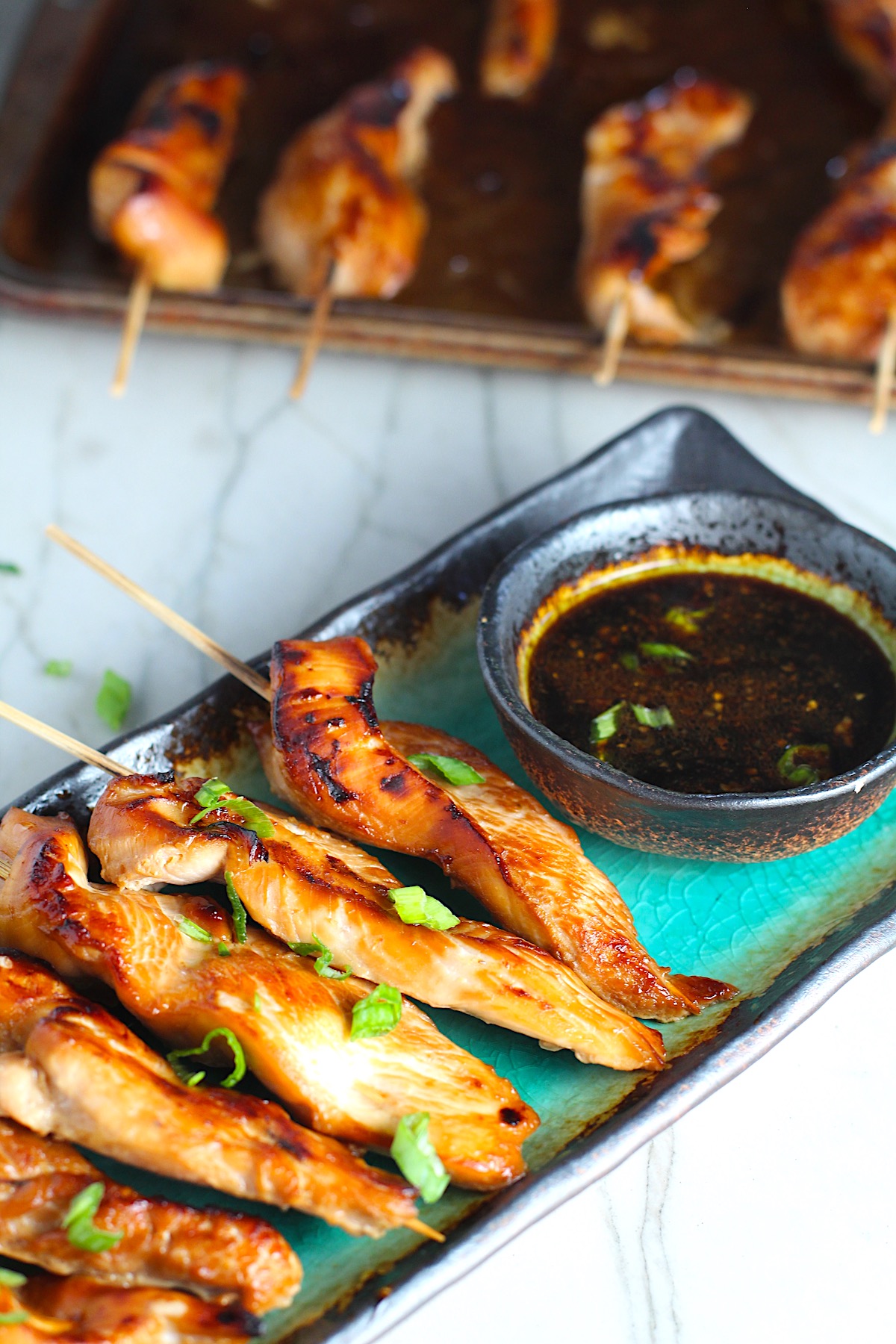 Chinese Chicken on a Stick Recipe on a rectangle platter with small bowl of dipping sauce and pan in background.