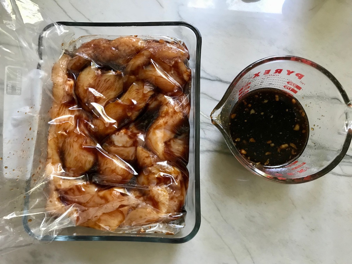 Raw chicken marinating in a storage bag in a glass container on counter with extra marinade in a measuring cup to the side for Chinese Chicken on a Stick Recipe.