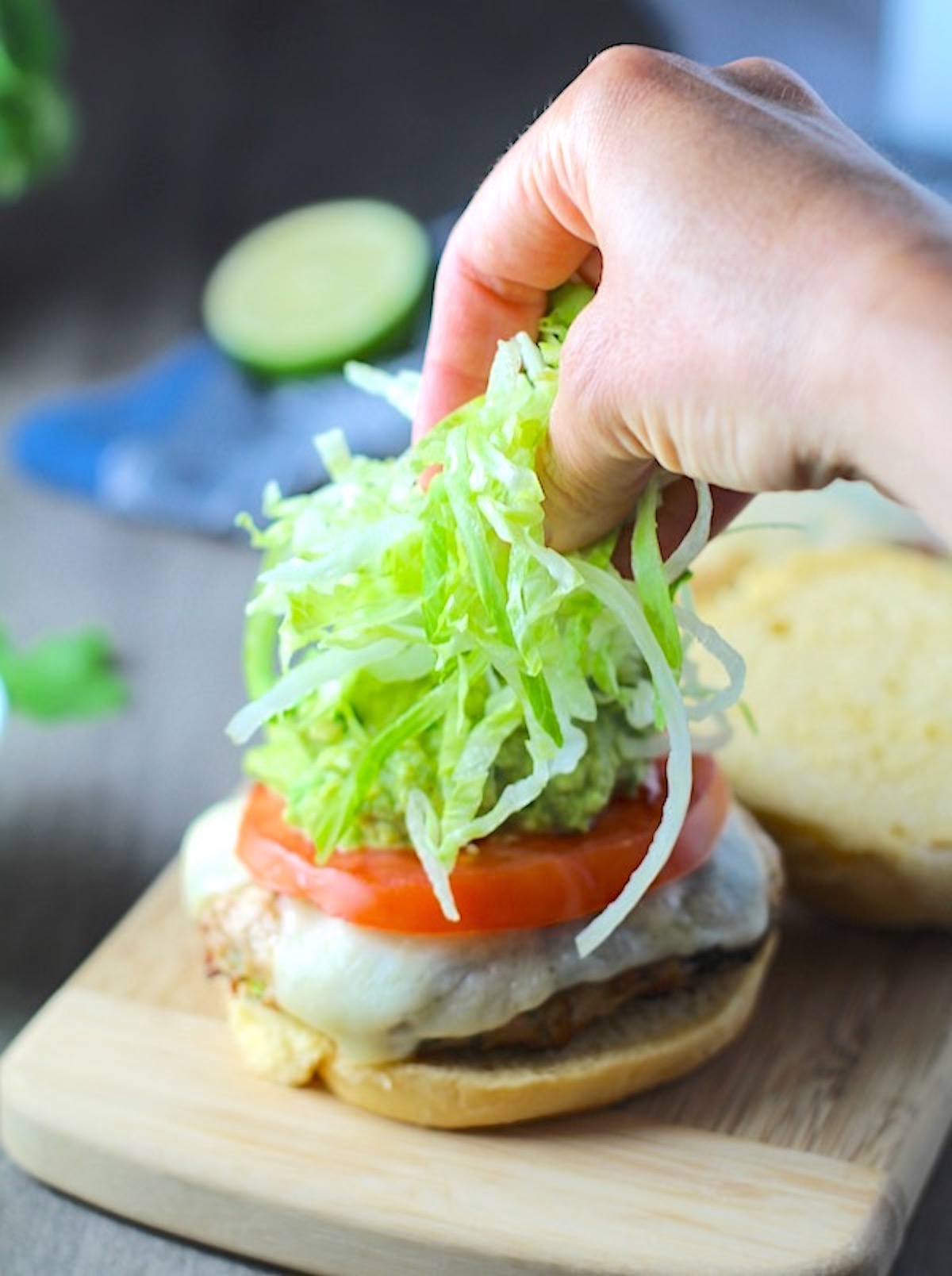 Hand putting lettuce on a Turkey Taco Burger with smokey taco seasonings, cilantro, and scallions, cheese, guacamole, tomato, and chipotle mayonnaise!