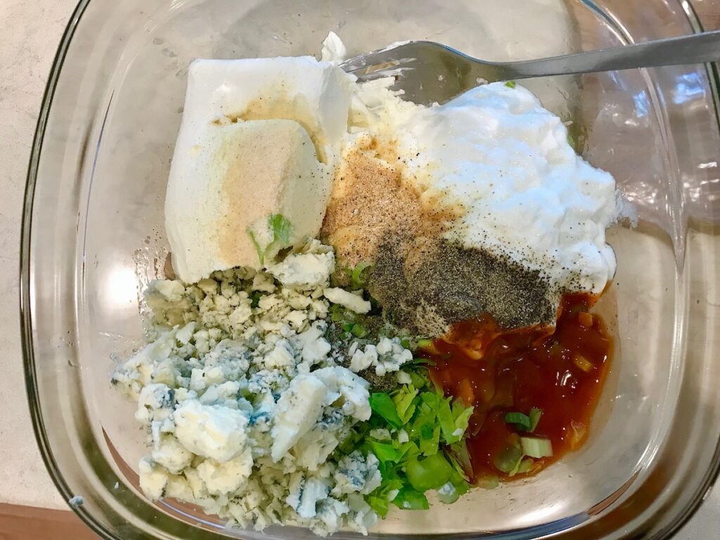 All ingredients added to a bowl for Buffalo Chicken Dip with greek yogurt. Ingredients are cream cheese, greek yogurt, shredded chicken, buffalo sauce, blue cheese, and scallions.