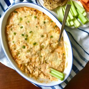 Buffalo Chicken Dip with greek yogurt in serving dish with spoon in it and a celery stick dipping into it. On the side is a plate of celery, carrots, and chips.