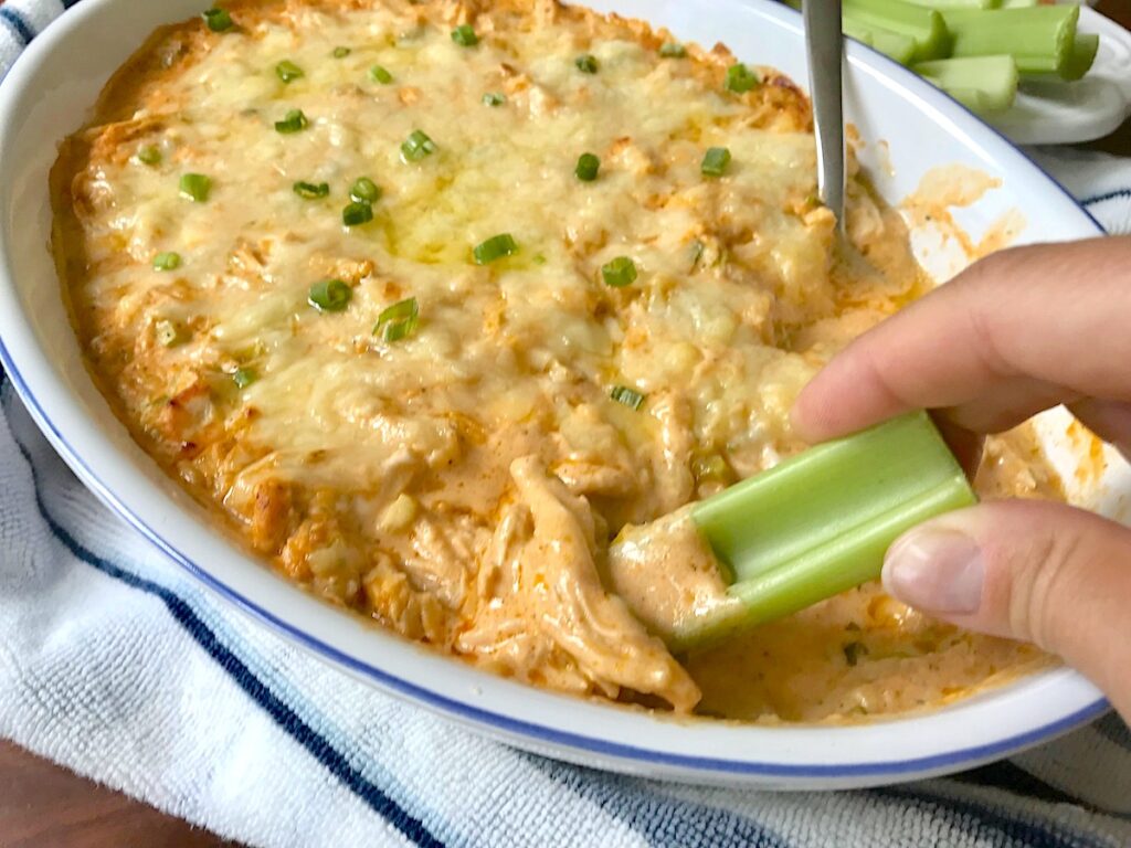 Hand with celery scooping Buffalo Chicken Dip with greek yogurt in serving dish with spoon in it.