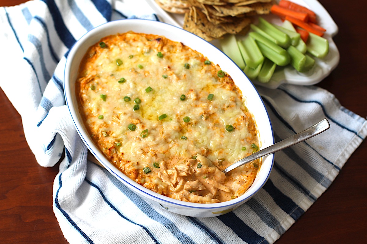 Buffalo Chicken Dip with greek yogurt in serving dish with spoon in it. On the side is a plate of celery, carrots, and chips.