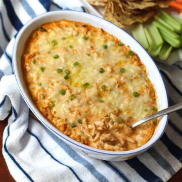 Buffalo Chicken Dip with greek yogurt in serving dish with spoon in it. On the side is a plate of celery, carrots, and chips.