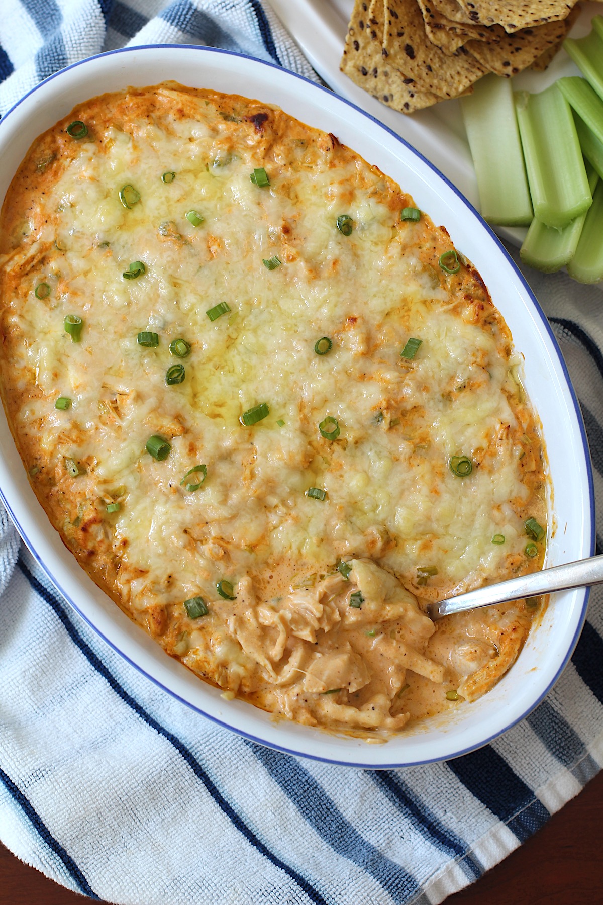 Buffalo Chicken Dip with greek yogurt in serving dish with spoon in it. On the side is a plate of celery, carrots, and chips.