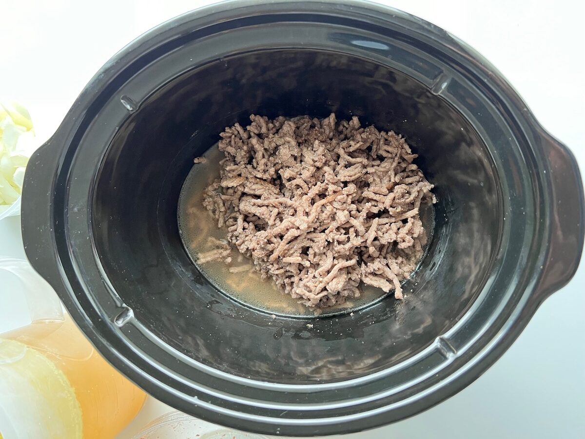 Cooked ground beef and liquid added to a slow cooker insert for Ground Beef Vegetable Soup Recipe. It's a perfect fall meal that's comforting, hearty, and delicious with loads of veggies!