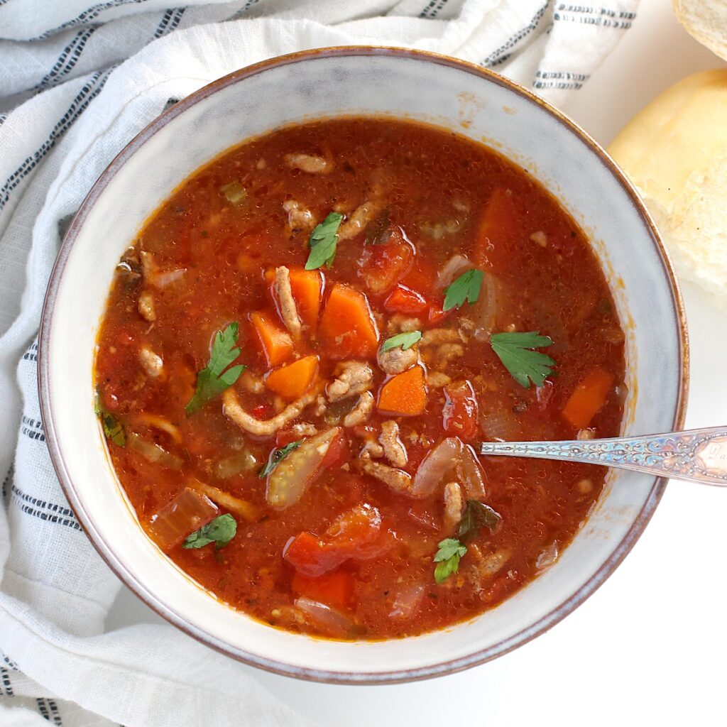 Ground Beef Vegetable Soup Recipe in a bowl with spoon on counter with blue and white towel around. It's a perfect fall meal that's comforting, hearty, and delicious with loads of veggies! Make it in the slow cooker or on the stove.