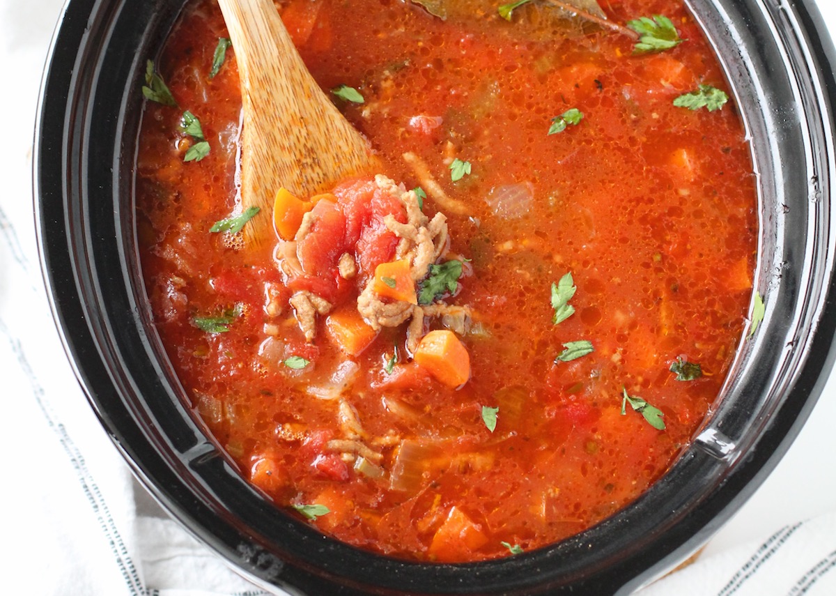 Ground Beef Vegetable Soup Recipe in a slow cooker pot with wooden spoon on counter with blue and white towel around. It's a perfect fall meal that's comforting, hearty, and delicious with loads of veggies! Make it in the slow cooker or on the stove.