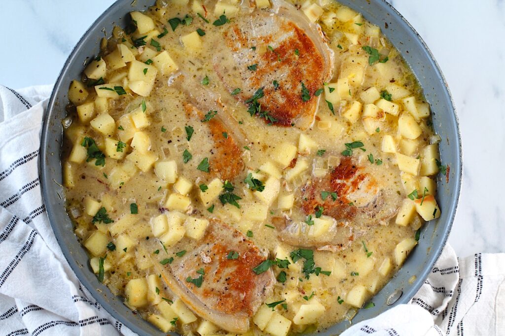 Pork Normandy in a skillet with a towel below on a counter. It has browned boneless pork chops with a savory sauce made of apples, onions, thyme, and cream.