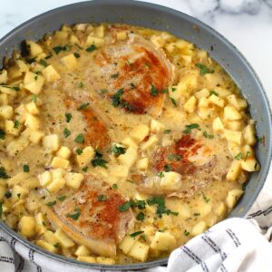 Pork Normandy in a skillet with a towel below on a counter. It has browned boneless pork chops with a savory sauce made of apples, onions, thyme, and cream.
