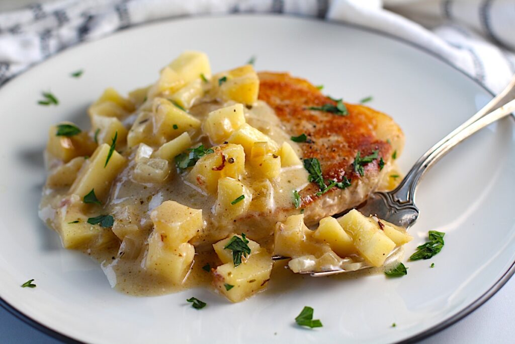 Pork Normandy on a plate with a fork and a towel below on a counter. It has browned boneless pork chops with a savory sauce made of apples, onions, thyme, and cream.