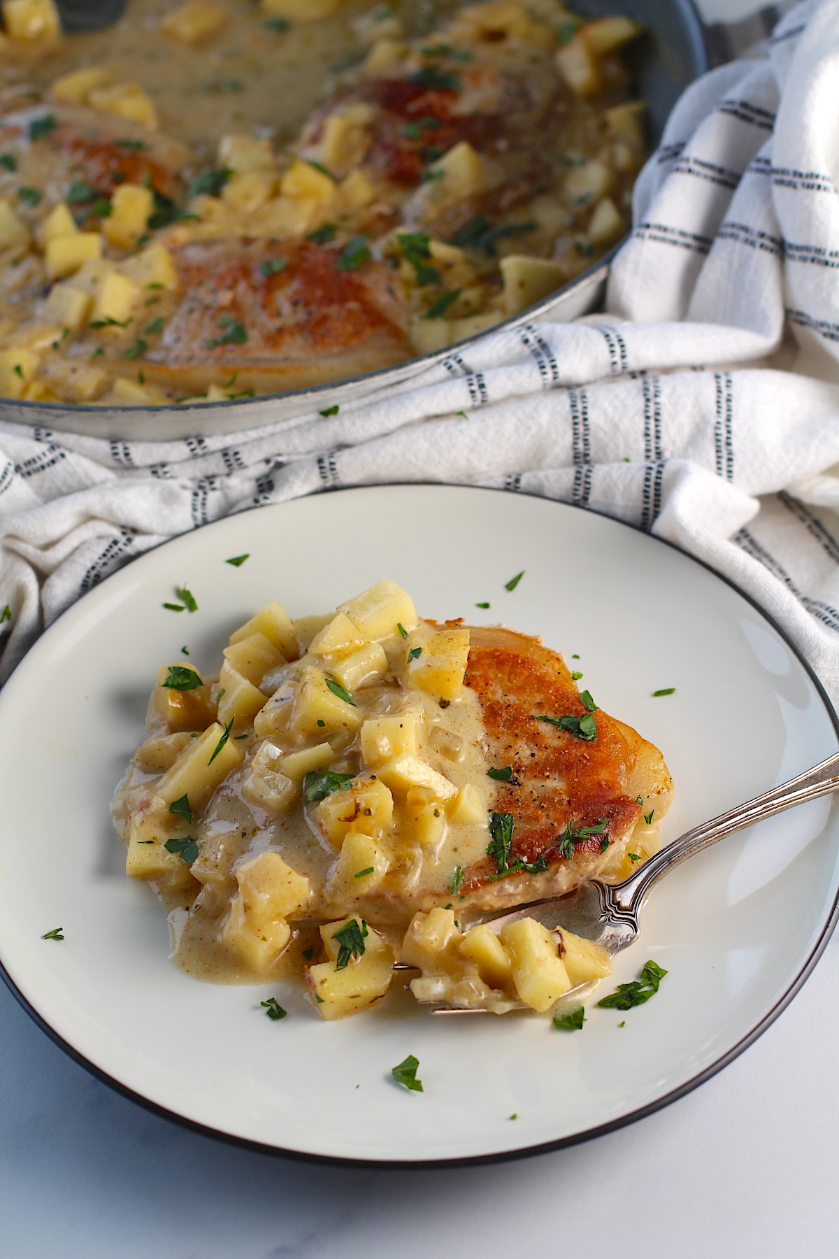 Pork Normandy on a plate with a fork and a towel below on a counter. It has browned boneless pork chops with a savory sauce made of apples, onions, thyme, and cream.