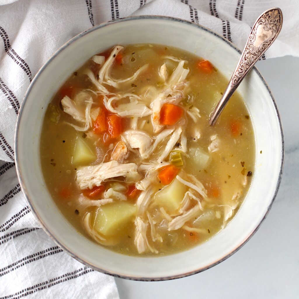 Creamy Chicken Potato Soup Recipe with carrots and celery in a bowl with a spoon on counter with towel. It's comforting, hearty, and delicious. It's a great dinner for busy families!