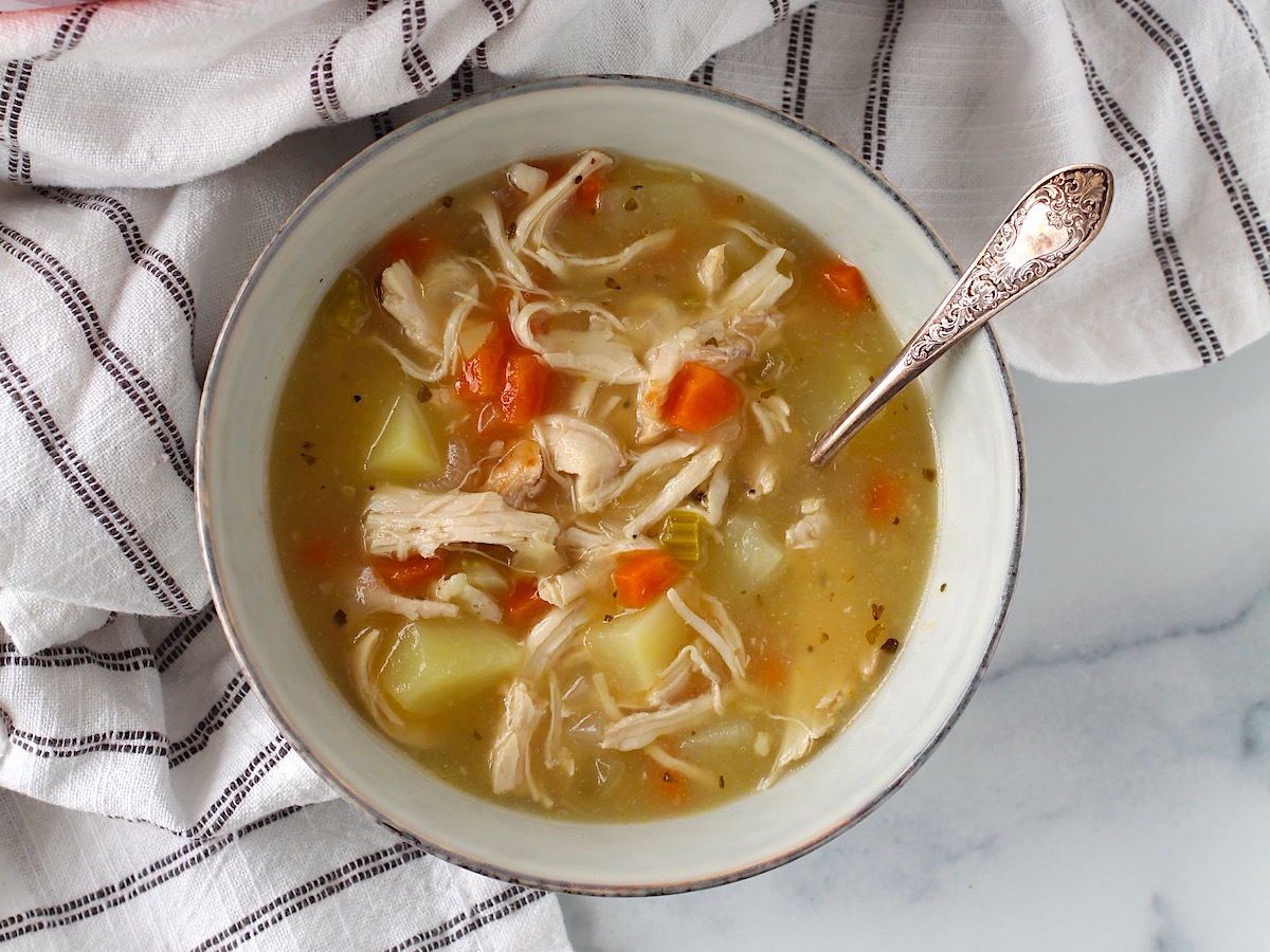 Creamy Chicken Potato Soup Recipe with carrots and celery in a bowl with a spoon on counter with towel. It's comforting, hearty, and delicious. It's a great dinner for busy families!