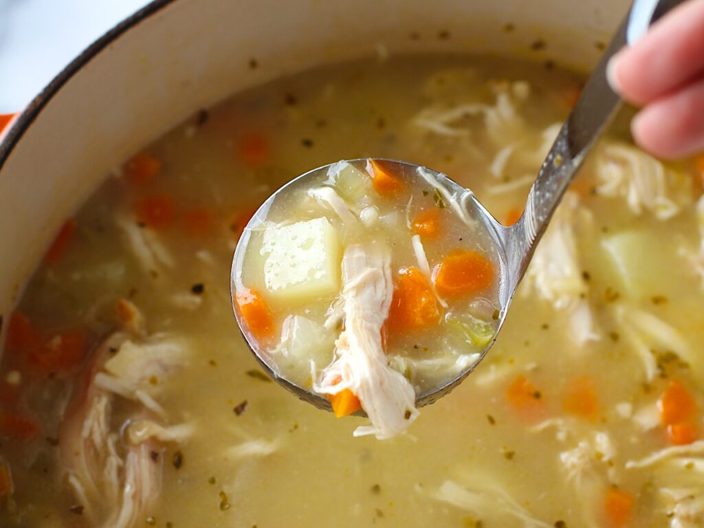 Hand holding ladle of Creamy Chicken Potato Soup Recipe with carrots and celery over a dutch oven filled with the soup on counter with towel. It's comforting, hearty, and delicious. It's a great dinner for busy families!