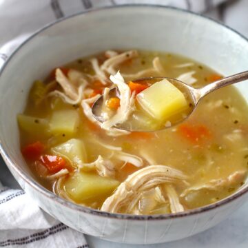 Hand holding spoon of Creamy Chicken Potato Soup Recipe with carrots and celery over a bowl filled with the soup on counter with towel. It's comforting, hearty, and delicious.  It's a great dinner for busy families!