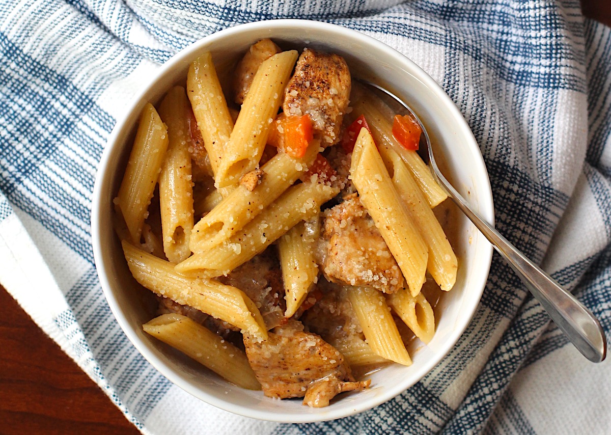 Bowl of Chipotle Chicken Pasta with penne pasta, diced red pepper, parmesan cheese sprinkled on top, and a fork in the bowl on top of kitchen towel.