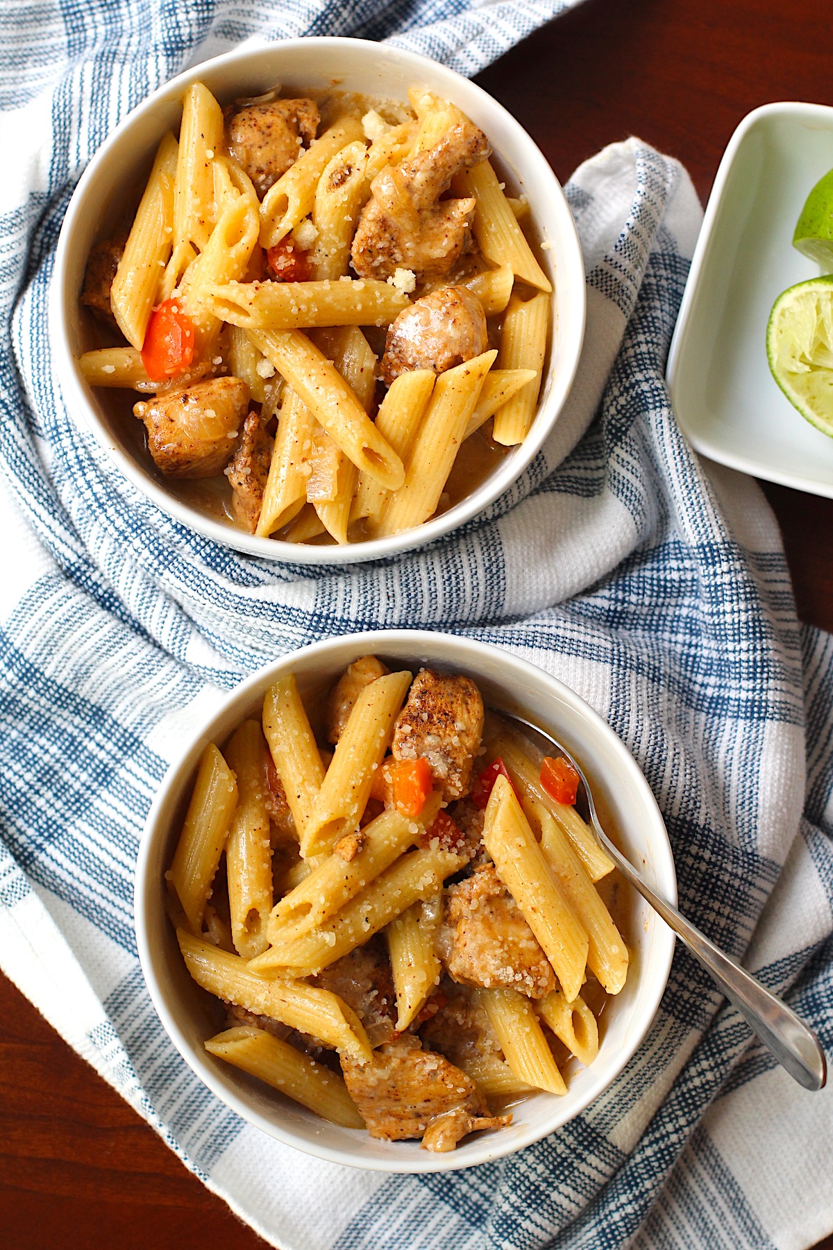 Two bowls of Chipotle Chicken Pasta with penne pasta, diced red pepper, parmesan cheese sprinkled on top, and a fork in the bowl on top of kitchen towel.