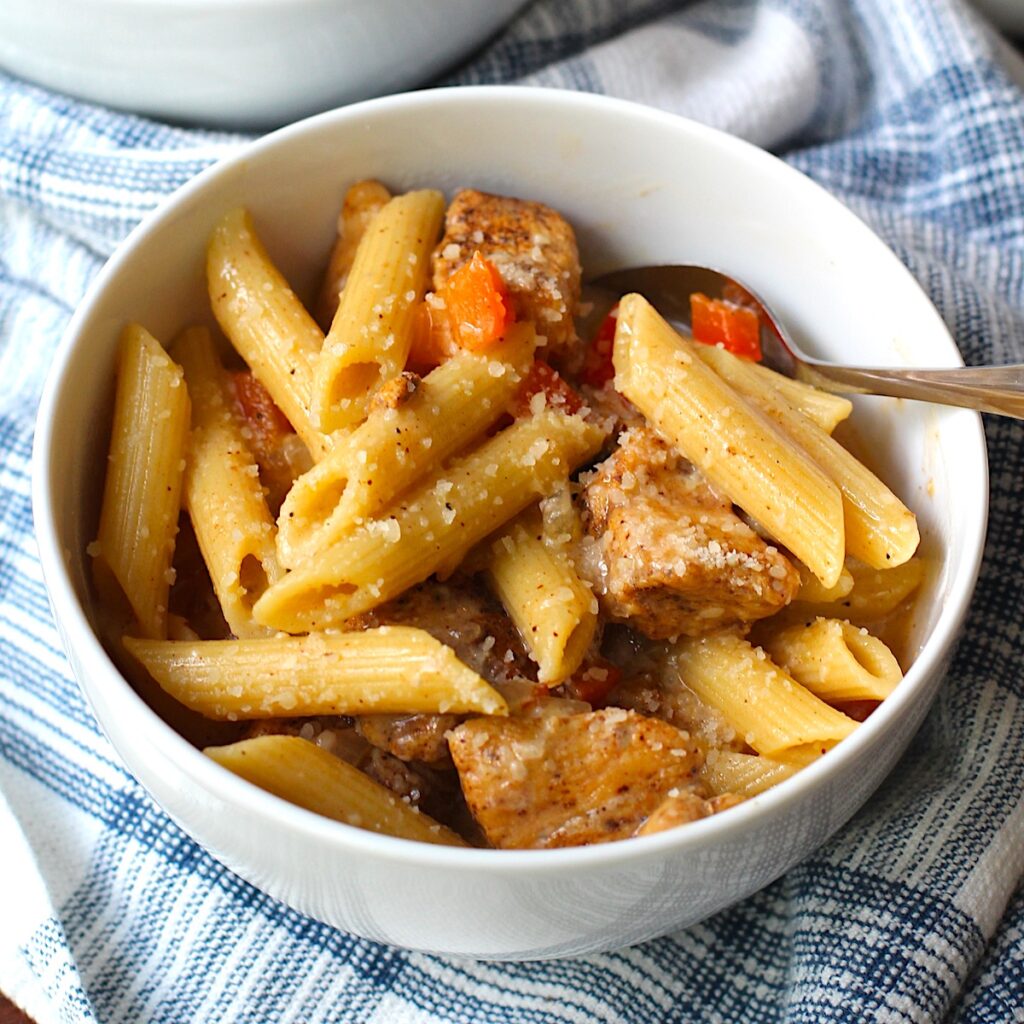 Bowl of Chipotle Chicken Pasta with penne pasta, diced red pepper, parmesan cheese sprinkled on top, and a fork in the bowl on top of kitchen towel.