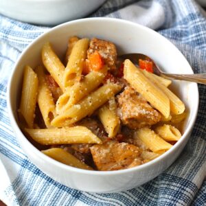 Bowl of Chipotle Chicken Pasta with penne pasta, diced red pepper, parmesan cheese sprinkled on top, and a fork in the bowl on top of kitchen towel.