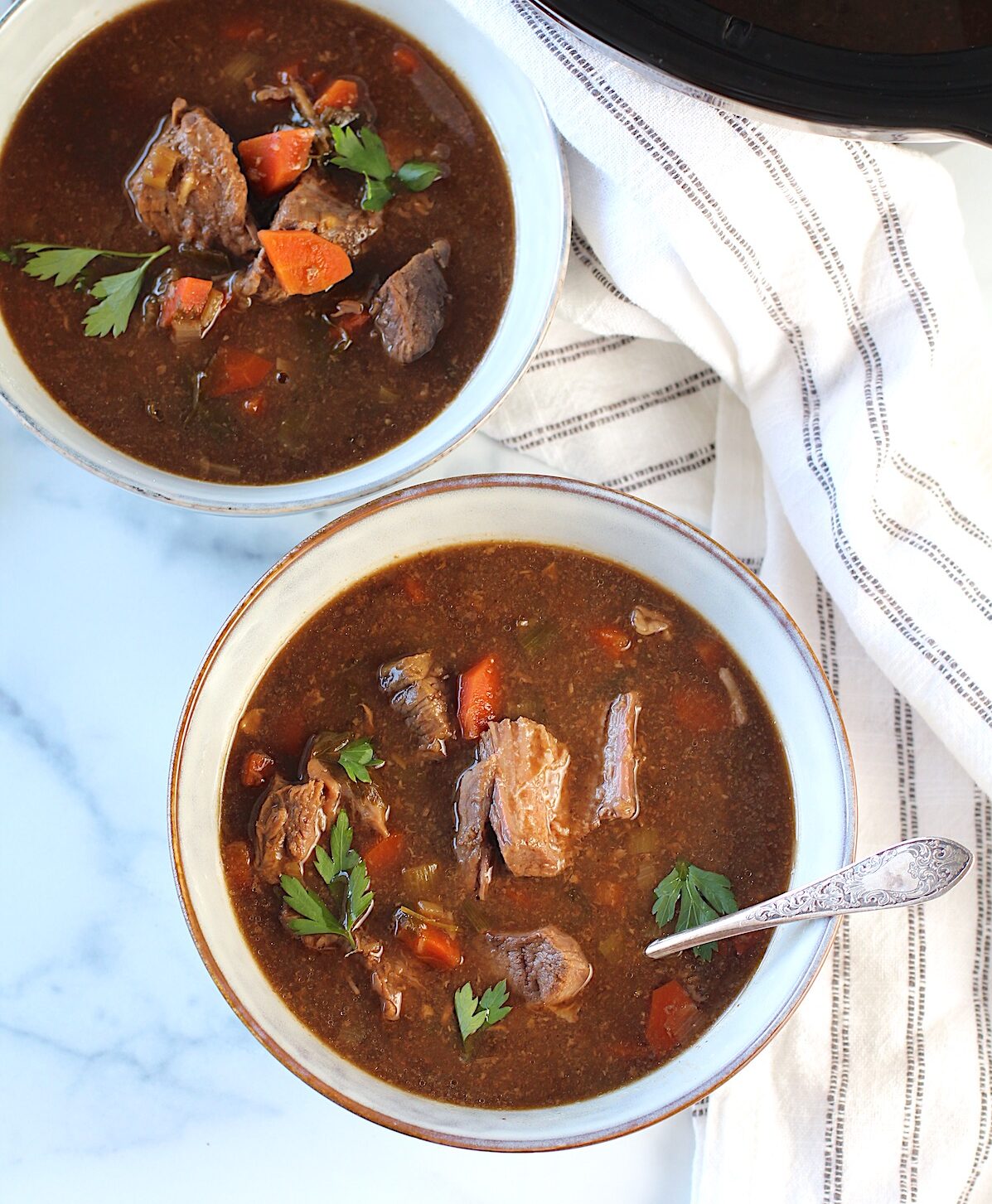 Two bowls of Korean Beef Stew, one with a spoon, on counter with blue and white striped towel. Korean Beef Stew is sweet, salty, tangy, and oh-so flavorful!