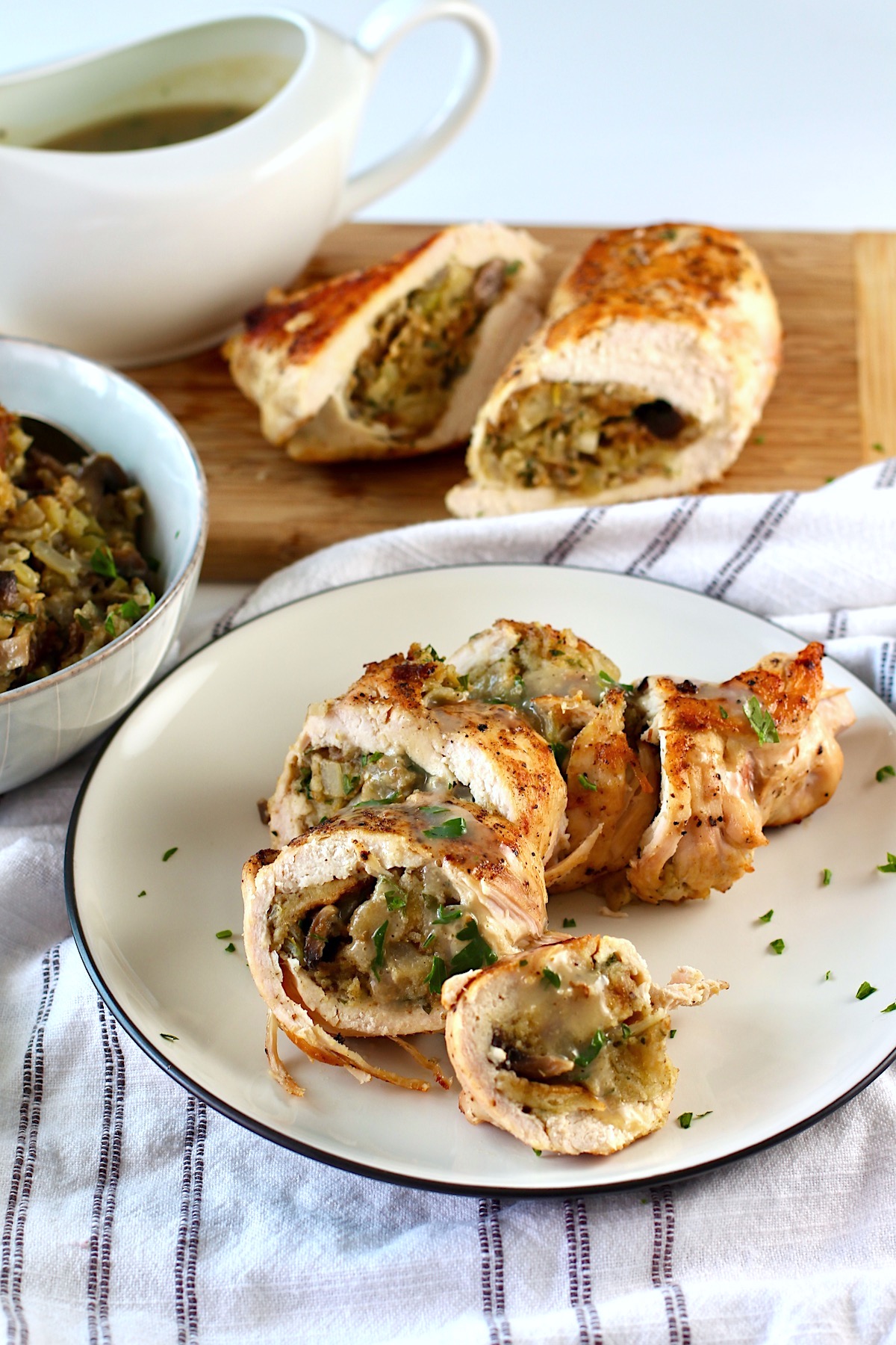 Stuffed chicken sliced on a place with another stuffed chicken breast cut in half in the background, a gravy boat in back and a bowl of stuffing to the side. Stuffing stuffed chicken breast recipes are easy and delicious!