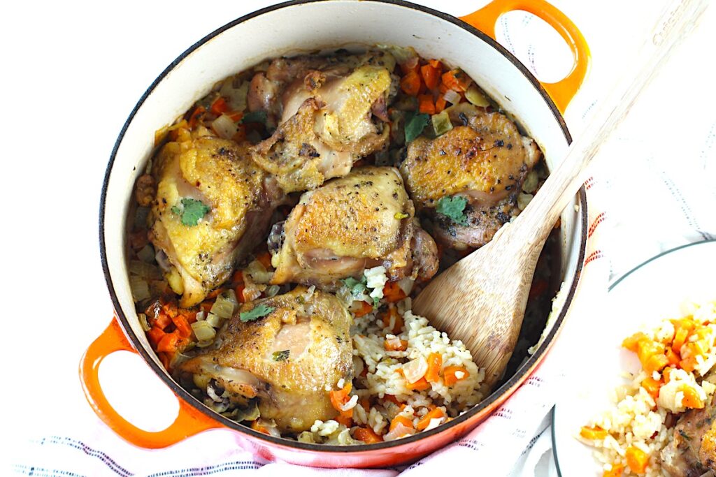 Roasted Chicken Rice in a pot with 5 crispy chicken thighs on top of rice with carrots and onions and a wood spatula scooping the rice. This recipe is an easy and delicious family dinner that you can make on any busy weeknight or cook entirely ahead!