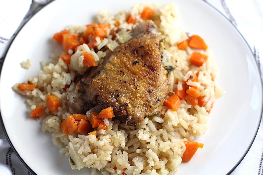Roasted Chicken Rice on a plate with one crispy chicken thigh on top of rice with carrots and onions. This recipe is an easy and delicious family dinner that you can make on any busy weeknight or cook entirely ahead!