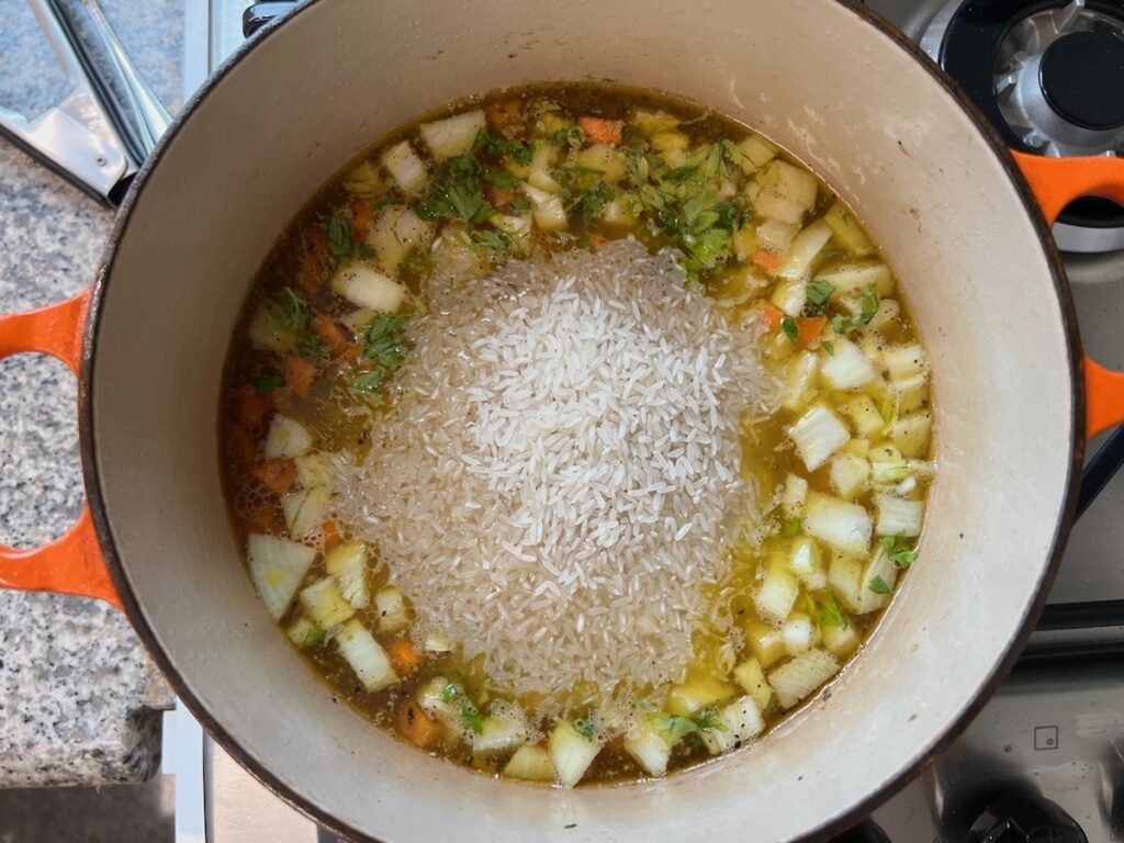 Rice added to carrots, onions, parsley, and broth cooking in a pot for Roasted chicken rice.