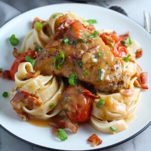 Smothered Chicken with Bacon, and Tomatoes and fettuccine on a plate with pan in background. The chicken is smothered in a thickened sauce infused with smokey and salty bacon flavor. Cherry tomatoes give a sweet and tangy pop.