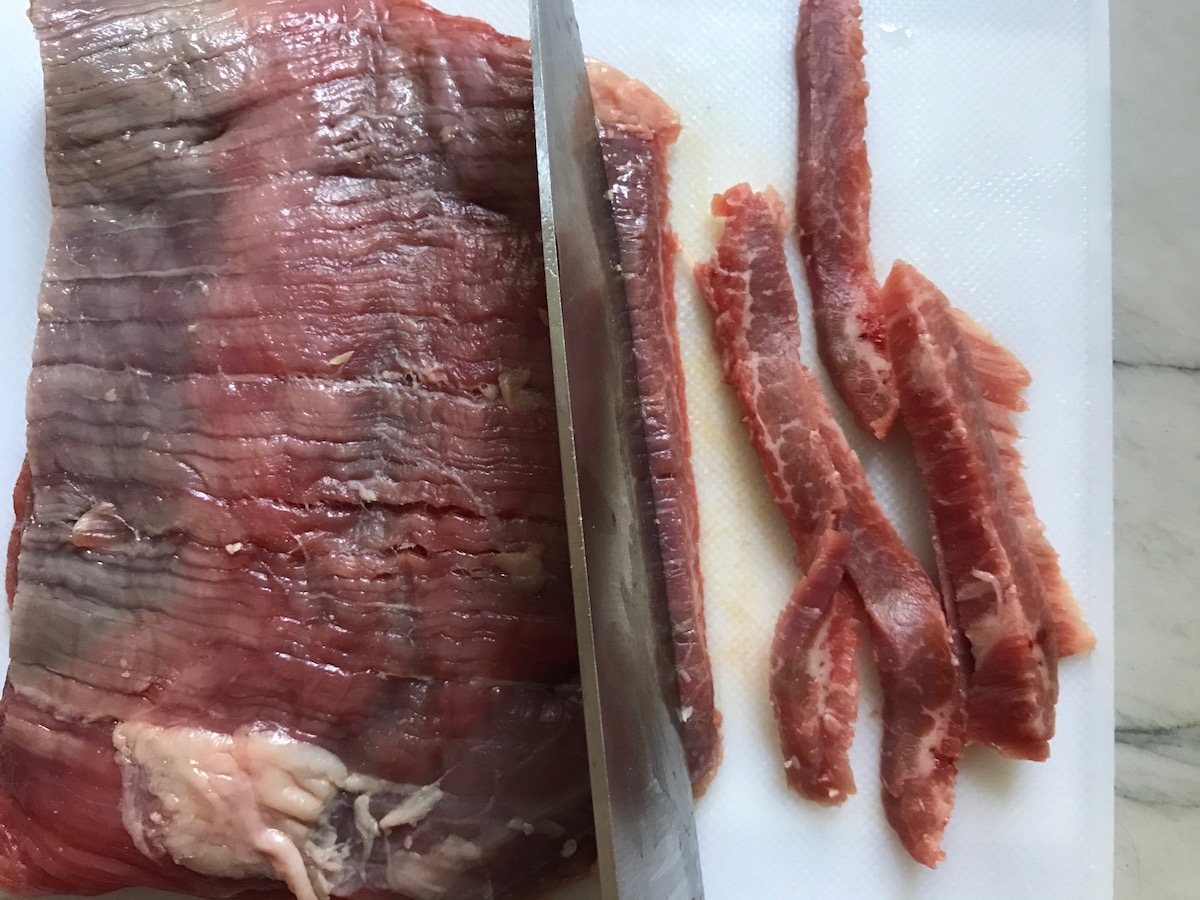 Knife cutting raw flank steak into strips against the grain for Philly Cheesesteak casserole recipe with Steak.