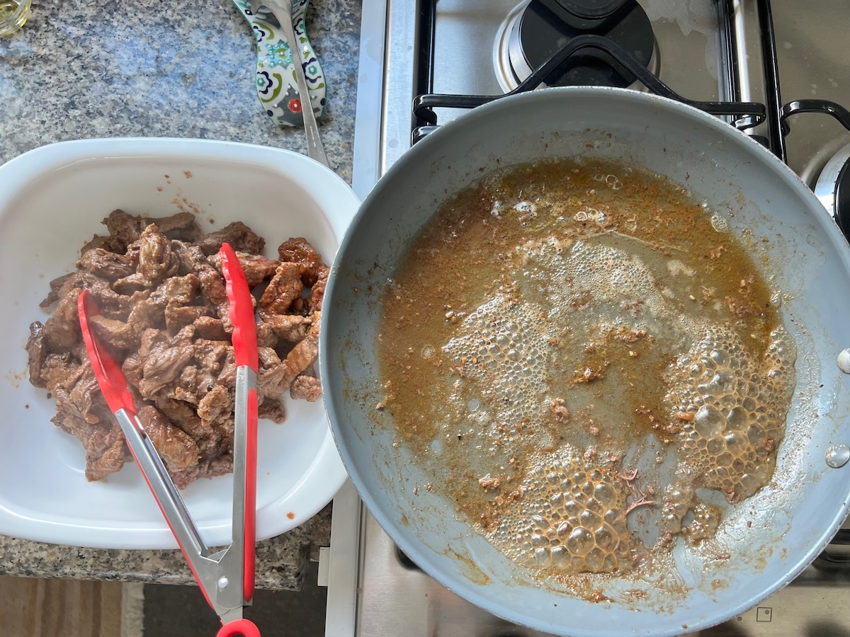 Tongs transferring cooked flank steak from skillet to casserole dish Philly Cheesesteak casserole recipe with Steak.