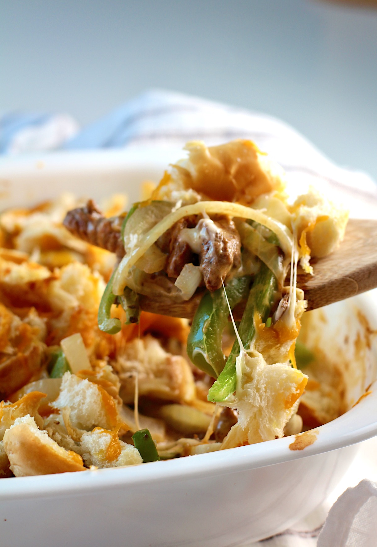 Wood spatula scooping up Philly Cheesesteak casserole recipe with Steak with peppers and onions cheesy toasted bread on top.