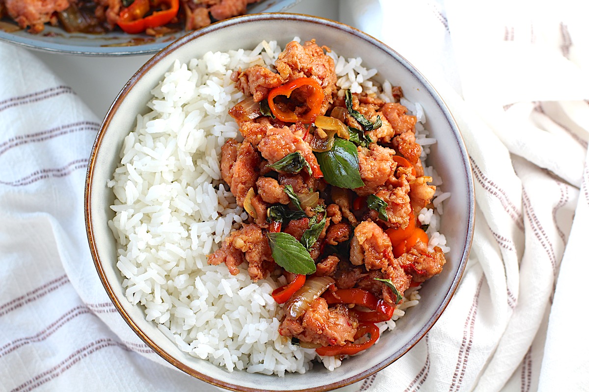 Thai Basil Pork Recipe with mini red pepper slices in a bowl over rice on the counter with a towel with skillet in background.
