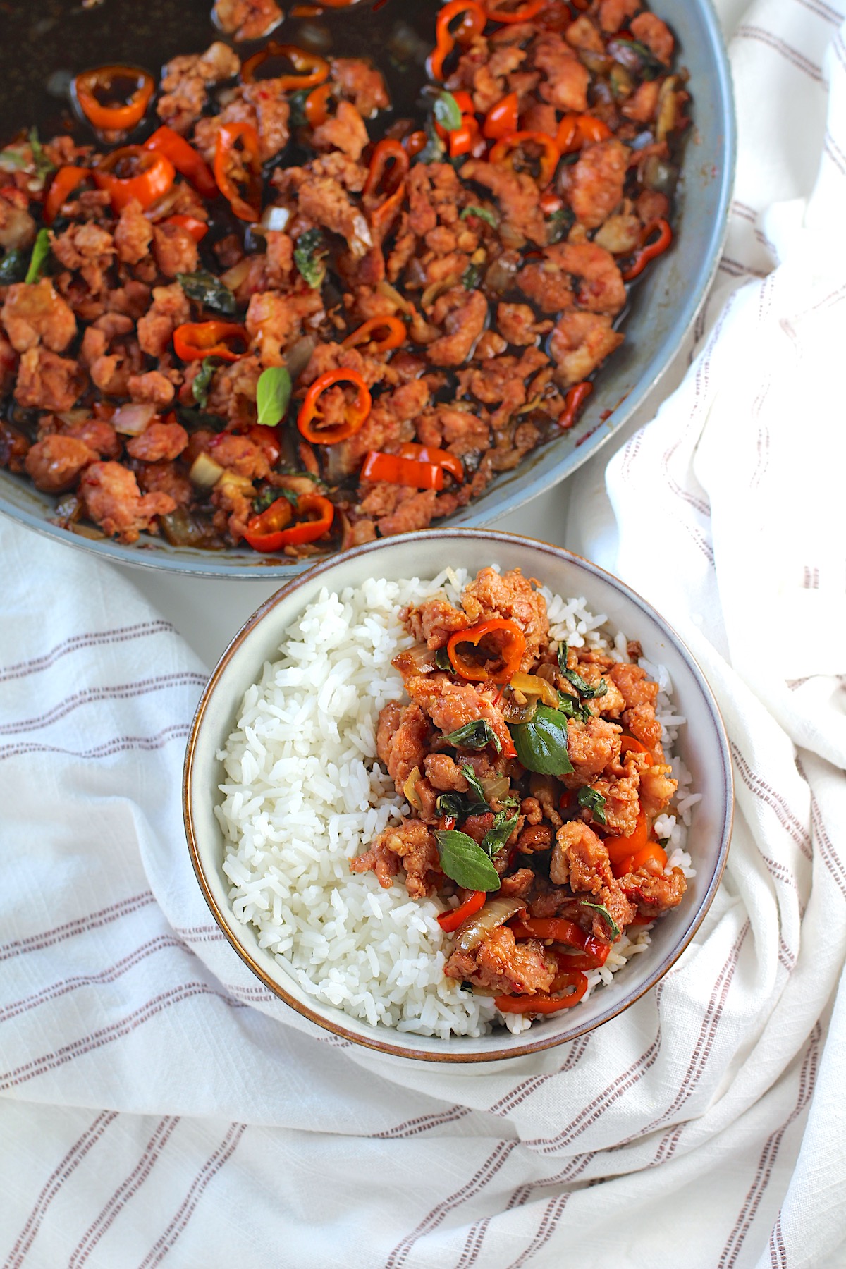 Thai Basil Pork Recipe with mini red pepper slices in a bowl over rice on the counter with a towel with skillet in background.
